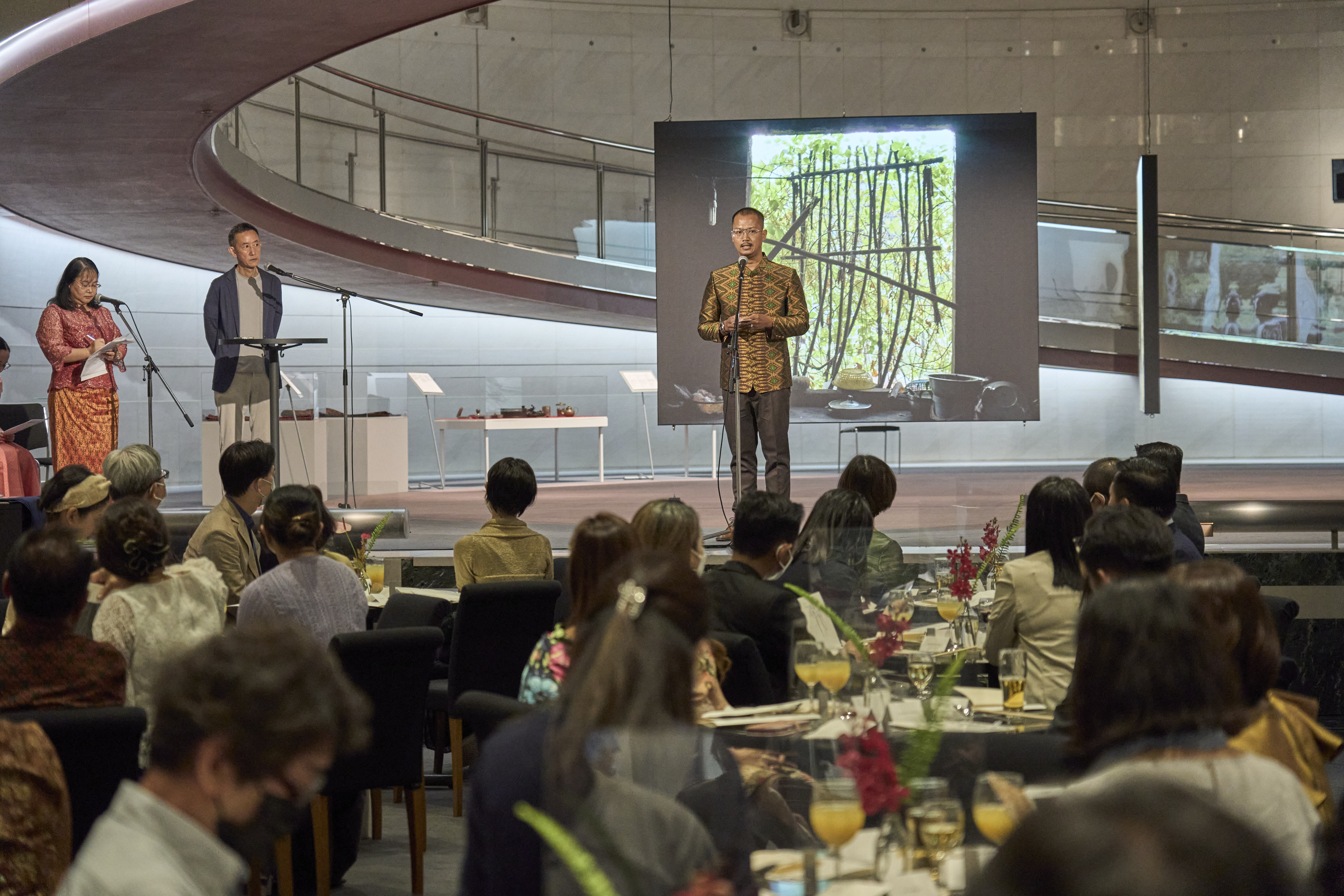 Slider Image Kim Hak speaks on a stage at his exhibition opening, in front of a crowd of attendees seated at tables