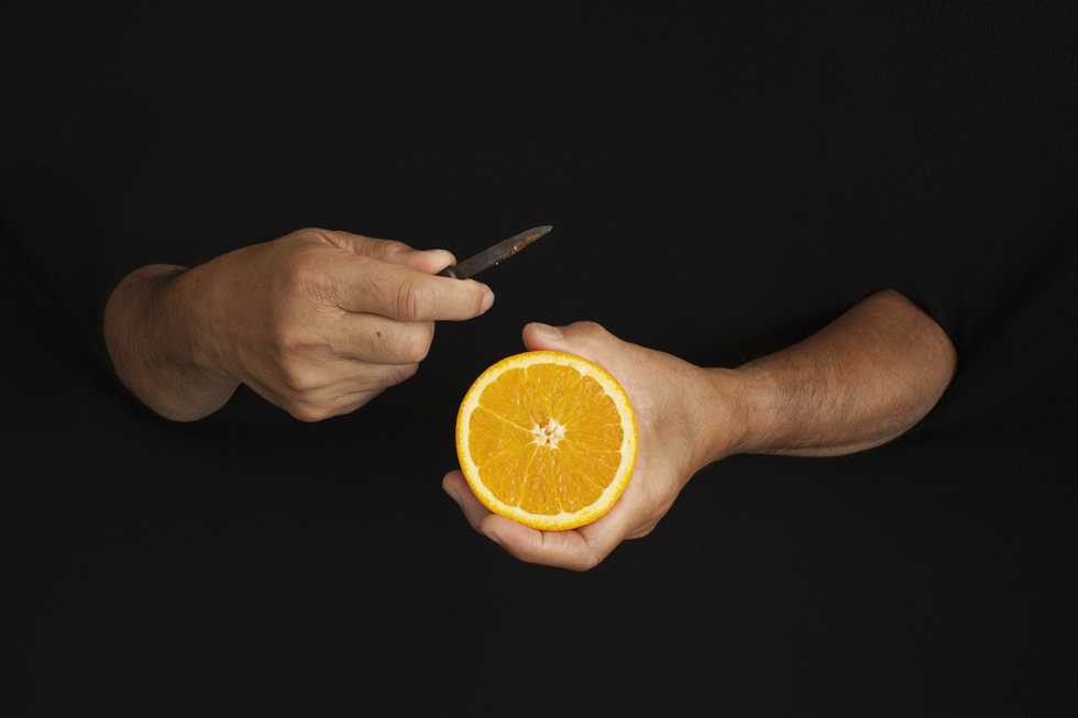Full Image Two visible hands, one holds half of an orange and the other holds a knife as if preparing to cut the orange