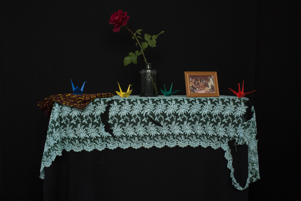 Full Image Kim Hak's photograph of a table, covered with a lace shawl, with three coloured origami cranes, a framed photograph and a single rose in a vase on top of it